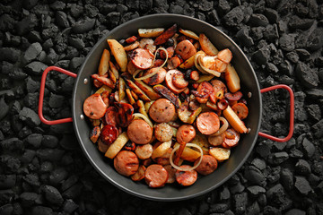 Frying pan with tasty potatoes and sausages on coals