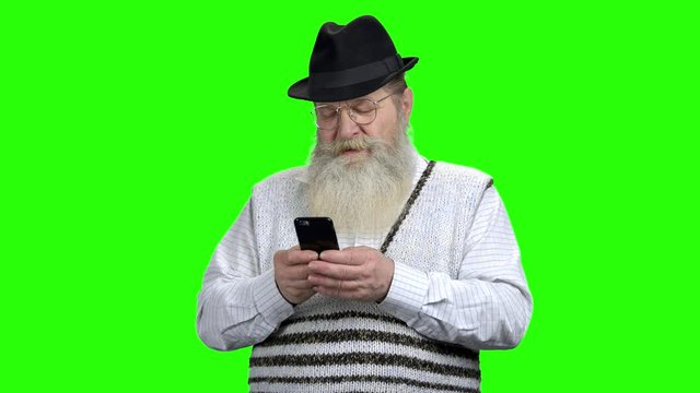 Senior man using mobile phone on green screen. Old bearded man in hat holding cell phone on Alpha Channel background.