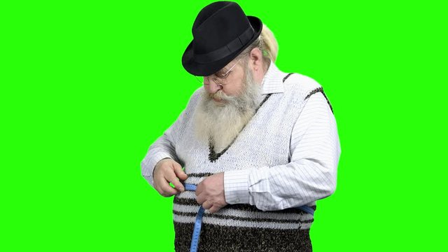 Elderly man measuring belly with tape measure. Displeased with result. Green Chroma Key background. Obesity and weight loss concept.