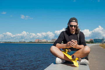 A young man sitting on a quay with a yellow skateboard on his lap is checking a smartphone.