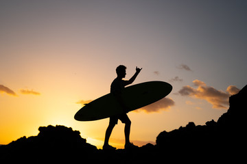 A male surfer at sunset comes with a surfboard over the cliffs of the ocean. Waiting for the wave. Silhouet of a sportsman guy in the rays of a summer warm sun. Shows hang loose or shaka sign.