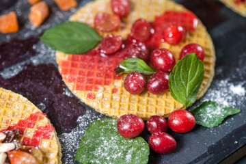 Fototapeta na wymiar Sweet dessert on dark background. Traditional breakfast with gaufre, waffle, crispy wafer with red cherry berries, mint, fruits and syrup.