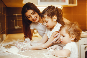 a young mother in a white t-shirt stands in the kitchen and works with flour along with her children