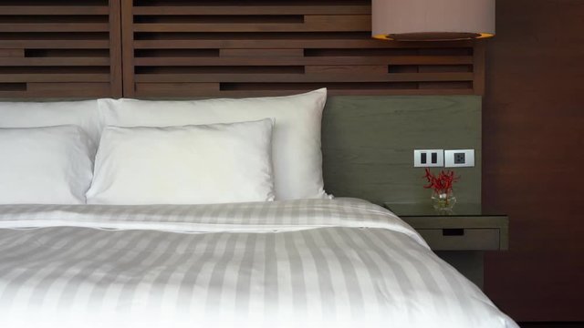 Pan across a made up completely white hotel bed with warm wood headboard.