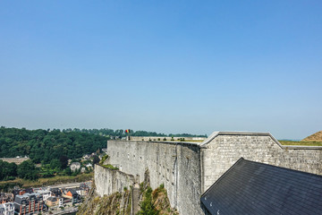 Dinant, Belgium - June 26, 2019: Gray stone south ramparts of Citadelle fortification with Belgian Flag at top under blue sky. Horizon is forest and down the Meuse River and cityscape.