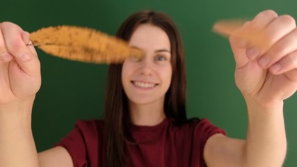 Portrait of young girl with long hair wearing red t-shirt, having fun playing with yellow autumn leaves and looking at camera, against green background. People and lifestyle.