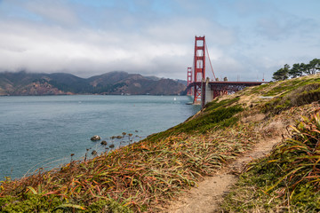 Fototapeta na wymiar Golden Gate Bridge with low clouds on the horizon and some scrubby plants and a worn dirt path in the foreground