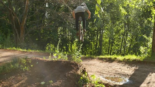Man cyclist pro rider jumping and flying dh mountain bike on springboard road gap in green forest. Bicyclist riding downhill on mountain bicycle. Extreme trick on mountain bike on forest trail