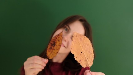 Cheerful young girl with long hair wearing red t-shirt, having fun playing with yellow autumn leaves and looking at camera, against green background. People and lifestyle.