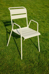 A beige metal chair stands on the lawn. Long hard shadows on the green grass. Urban theme. Theme of loneliness