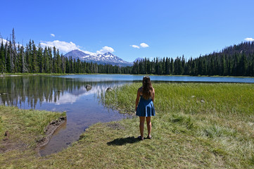 Fototapeta na wymiar Young woman enjoys view of Middle and North Sisters volcanoes reflected in Scott Lake, Oregon on a calm sunny summer afternoon.