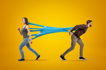 Fototapeta na wymiar Young brunette girl wearing casual jeans and t-shirt stuck to young man in casual clothes with blue sticky slime on yellow background.