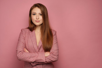 Red-haired girl in a business suit stands on a pink background with her arms folded and smiling looking at the camera.