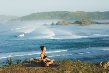 Woman practicing yoga and sitting in lotus pose outdoor with amazing ocean view. Health and well-being concept. nature background.