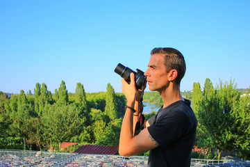 Man in black T-shirt taking pictures with a camera in profil. In nature with vegetation and countryside in the background. © Bruno