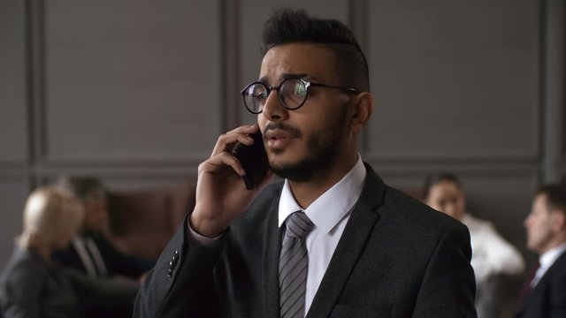 Chest-up shot of frowning frustrated 30-something Middle Eastern businessman in suit, tie and glasses calling and starting to talk on phone, while colleagues are sitting around table in background