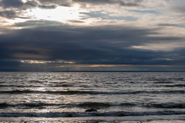 Bad weather sea with a boat with a sail on the horizon. The sun through the dark clouds in the sky. Small waves.