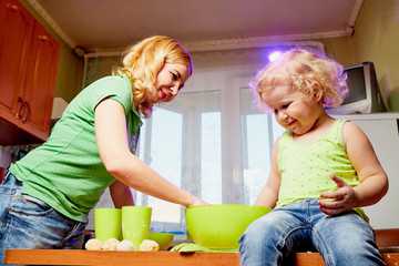 Mother and her three years old blonde daughter are cooking in a kitchen. Happy mom and small girl with the dough