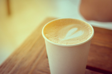Close up white paper cup of hot coffee (cappuccino, latte) with heart shape milk foam topping on wooden table with flare light and copy space for your text. Vintage tone.