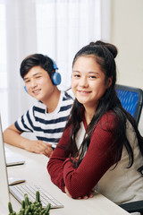 Smiling mixed-race pretty teenage girl sitting in front of computer at her desk, her cheerful brother listening to music in background