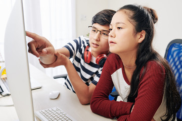 Smart teenage boy in glasses explaining part of programming code to his classmate when they are...