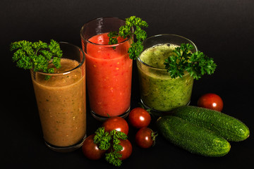 Still life. Vegetable juices in glass glasses, tomatoes, yellow pepper, cucumbers, green leaves of parsley on a black background. Close-up. Isolated. Vegetarianism.