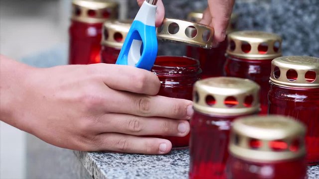 Man opens the lid and lights grave candle in red jar with lighter, hands closeup. Commemorate for victims, fallen people and fallen soldiers on the monument memorial steps.