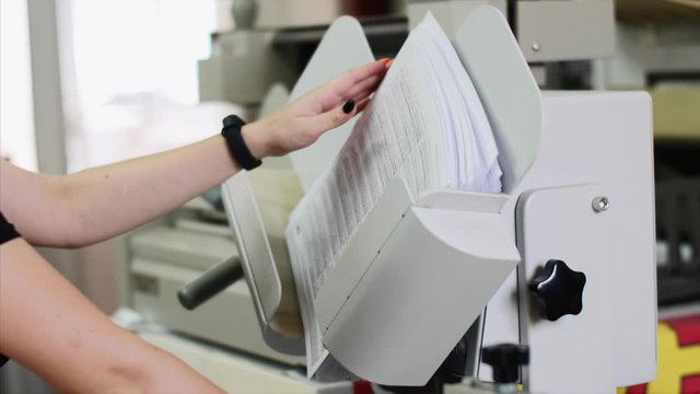 Woman is using shaking machine to straighten sheets of paper in printing house, part of manufacturing process, hands closeup. Alignment of stack of paper documents in printing house in shaking folder.