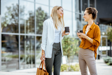 Two young businesswomen talking near the office building, having a small talk during the coffee...