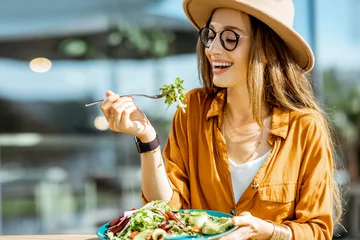 Fotobehang Stijlvolle jonge vrouw die gezonde salade eet op het terras van een restaurant en zich gelukkig voelt op een zomerdag © rh2010