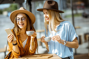 Two female best friends spending time together on the cafe terrace, feeling happy standing with coffee and phone during a summer day
