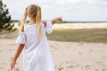 Fototapeta na wymiar Young girl with long blond hair in the white dress points to something outdoors 