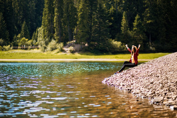 Freedom. Happy young woman sitting on beach of forest lake with arms raised, outdoor.