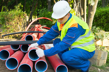 builder, in a white helmet, pulls out red plastic pipes into a trench, for laying an electric cable...