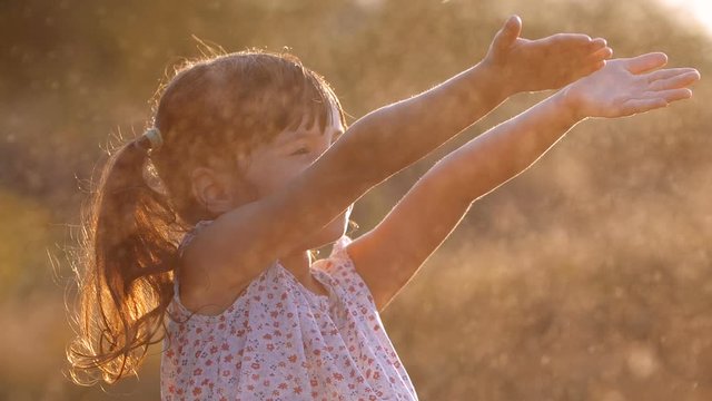 Close up of little cute girl  four years old having fun joyfully reaches out to flying drops of water and catching them in warm bright sunset rays on sunny summer evening. Slow motion 200 fps 