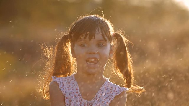 Close up of little cute girl four years old having fun joyfully jumping in flying spray of water, funny showing tongue in warm bright sunset rays on sunny summer evening. Slow motion  200 fps 
