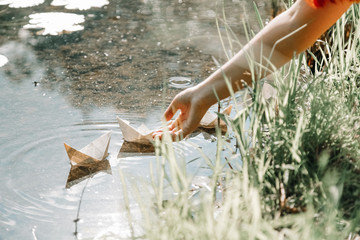 Cheerful girl do paper boats near the river in the sunny day city park alone