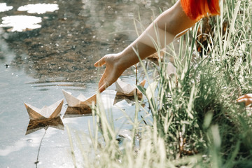 Cheerful girl do paper boats near the river in the sunny day city park alone
