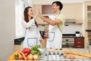 Happy young couple standing at kitchen table in aprons and giving each other high five