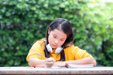 Little Asian schoolgirl doing her homework in the park with headphones on her neck.