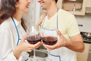 Positive young couple looking at each other and toasting with glasses of wine when standing in kitchen in aprons