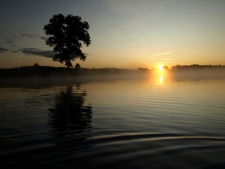 Fototapeta premium foggy blured orange sunrise at a river, the burning sky is reflected in calm water , dark tree silhouettes, Salaca river, Burtnieks lake, Latvia