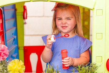 A cute blonde little girl with a funny face, blowing soap bubbles inside a colorful toy house