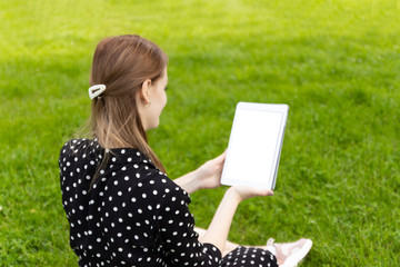 Young blonde girl in a summer dress makes selfie on a tablet while sitting on green grass in a park.