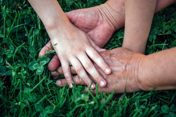 Hands unrecognizable grandmother and her granddaughter on green grass. Hands of elderly woman and young girl, close-up. Age and generational difference. Aged and wrinkled hands with young hands.