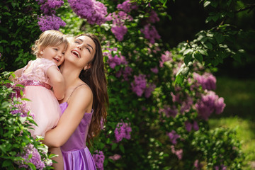 Laughing woman with kid girl on a walk. Blooming lilac trees on background