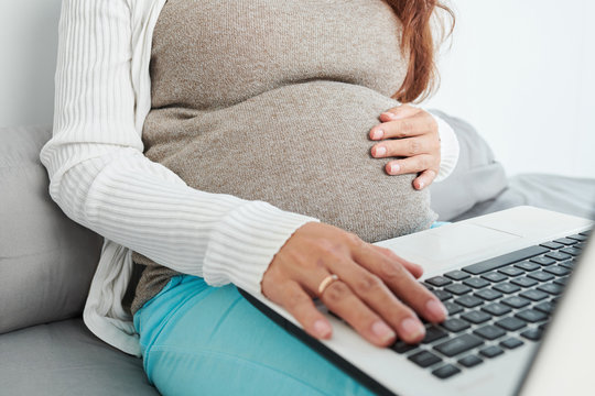 Close-up image of pregnant woman touching her baby kicking in womb and searching for information on laptop