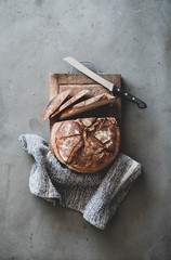 Flat-lay of freshly baked sourdough bread loaf and bread slices on rustic wooden board over grey concrete table background, top view, vertical composition