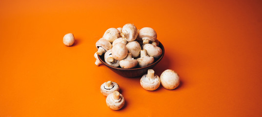 Mushrooms in a wooden bowl on an orange background. The small white champignon in a plate and scattered near it.