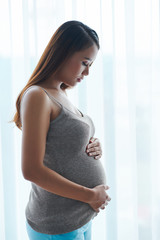 Calm young Vietnamese pregnant woman standing against window and touching her belly